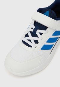 Pantof sportiv alb cu accente albastre, partea superioară din plasă și talpă de cauciuc. Dispune de o curea cu velcro și șireturi tradiționale pentru suport.