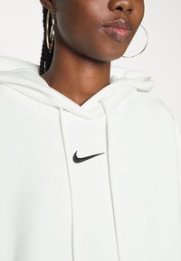 Hvit hettegenser laget av mykt stoff med en frontlomme, med en svart Nike-logo. Designet inkluderer en hette med snøring og ribbestrikkede mansjetter.