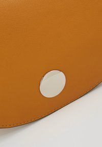 Torba ze skóry w kolorze jasnobrązowym o gładkiej fakturze i zaokrąglonym kształcie, z detalem w postaci srebrnego metalowego guzika oraz widocznym przeszyciem wzdłuż krawędzi.