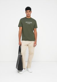 Olijfgroen katoen t-shirt met witte print "GUESS EST. 1981", gecombineerd met beige broek en een zwart tote bag, gedragen met witte sneakers.