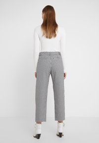 Donna con capelli lunghi, che indossa una maglietta bianca a maniche lunghe, pantaloni a scacchi bianco e nero e stivaletti bianchi, di spalle.