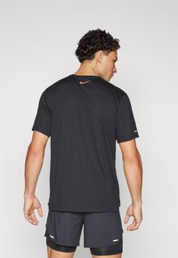 Svart treningst-skjorte med korte ermer, med en oransje Nike-logo ved bakhalsen, kombinert med grå shorts. Slett tekstur.