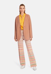 Hellrosa übergroße Blazer über einer gelben Bluse, kombiniert mit weit geschnittenen, gemusterten Hosen mit Chevron-Designs in Pink, Grün und Orange.