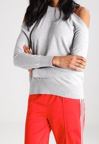 Pull en tricot gris avec un design à épaules dénudées, col rond et poignets côtelés. Associé à un pantalon de jogging rouge avec des rayures blanches.