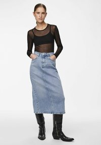 Haut en voile noir à manches longues associé à une jupe maxi en denim bleu clair. Le modèle porte des bottes noires jusqu'aux genoux, les mains dans les poches. Design simple et épuré.