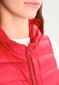 Röd puffy väst med hög krage, med frontdragkedja och quiltad textur. Bärs över en vit skjorta, detaljer av sömnad synliga.