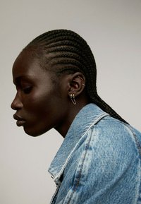 Chaqueta de denim con un lavado azul claro, cabello trenzado recogido, con pendientes dorados y sutiles luces en la piel. Perfil lateral.