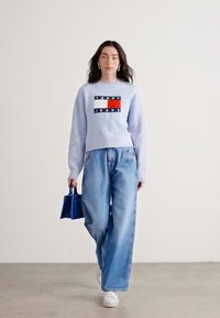 Lichtblauwe geribbelde trui met logopatch, gecombineerd met wijde blauwe jeans en witte sneakers, en een kleine blauwe handtas in de hand.