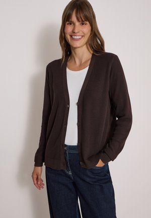 Donna sorridente che indossa un cardigan marrone scuro con bottoni, una camicia bianca e jeans blu, con una mano in tasca su uno sfondo semplice.