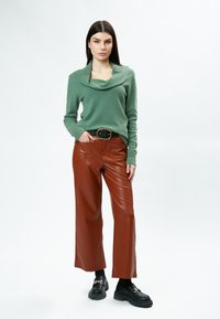 Grüner Strickpullover mit weitem Kragen, kombiniert mit braunen weit geschnittenen Hosen aus Kunstleder und schwarzen Plateau-Schuhen mit goldenen Besätzen.