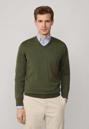 Olivgrüner V-Ausschnitt-Pullover aus weichem Material, mit langen Ärmeln und geripptem Saum. Über ein hellblau gestreiftes Hemd getragen.