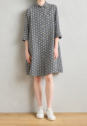 Person iført en knælængde knapklædt kjole med sort-hvide geometriske mønstre og hvide sneakers, stående mod en ensfarvet væg.