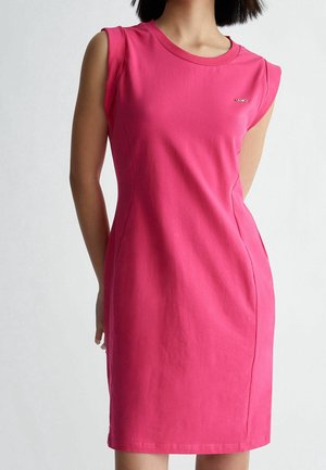Frau trägt ein eng anliegendes, ärmelloses, leuchtend pinkes Kleid mit rundem Ausschnitt und kleinem Metalllogo auf der Brust, steht vor einem schlichten Hintergrund.