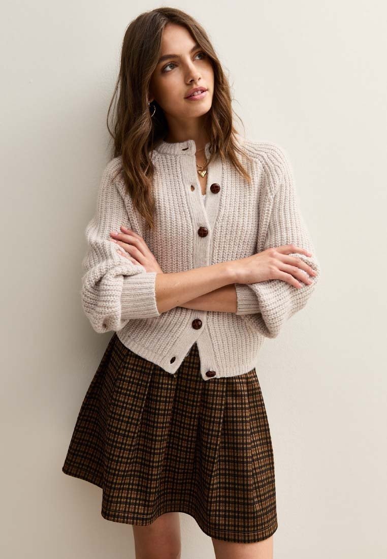 Cardigan spesso beige lavorato a maglia con bottoni in legno, indossato sopra una gonna a quadri marrone. Il cardigan presenta un colletto alto e maniche ripiegate.