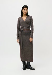 Kvinde iført en brun omslagsstriktrøje med en matchende lang nederdel og sorte ankle boots, stående mod en hvid baggrund.