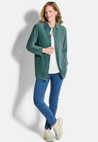 Grüne, knöpfbare Shirtjacke mit Kragen und Brusttasche, über einem weißen Shirt getragen. Kombiniert mit blauen Jeans und hellen Sneakers.