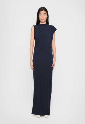 Woman standing facing forward wearing a sleeveless navy blue long dress with asymmetrical shoulder design and white pointed shoes.