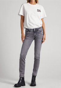 Valkoinen puuvillapaita, jossa on musta "Pepe Jeans" -logo, yhdistettynä istuviin harmaisiin farkkuihin ja mustiin nahkaisiiin nilkkabootseihin.