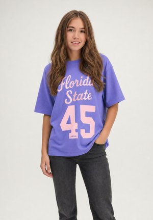 Jonge vrouw met lang bruin haar draagt een paarse "Florida State 45" T-shirt en zwarte spijkerbroek, staand met één hand in de zak.