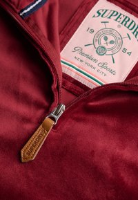 Vêtement rouge à fermeture éclair avec une texture lisse ; doté d'une fermeture éclair argentée et d'une tirette en cuir marron. L'étiquette affiche "Superdry" ainsi que des motifs liés au sport.