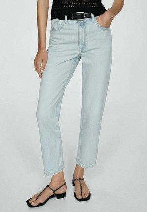 Nainen pukeutuneena vaaleansinisiin suoralahkeisiin farkkuihin, mustaan vyöhön, mustaan hihattomaan toppiin ja mustiin remmikenkiin, seisoo käsi yhdessä taskussa.