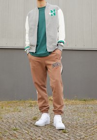 Giacca bomber grigia con maniche bianche e dettagli verdi, abbinata a pantaloni jogger marroni con logo "NIKE" e sneakers bianche.