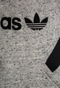Šedý svetr z materiálu s tečkovanou texturou. Na přední straně se nachází velký černý nápis "adidas" a černé logo se třemi pruhy.