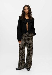 Femme debout portant un cardigan noir ouvert avec un grand col et un pantalon large en imprimé zèbre marron, avec des chaussures noires, sur fond blanc.
