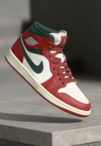 Crvena, bijela i zelena Air Jordan 1 tenisica s Nike swoosh oznakom, vezice zavezane, lebdi iznad betonske ivice protiv tamne pozadine.