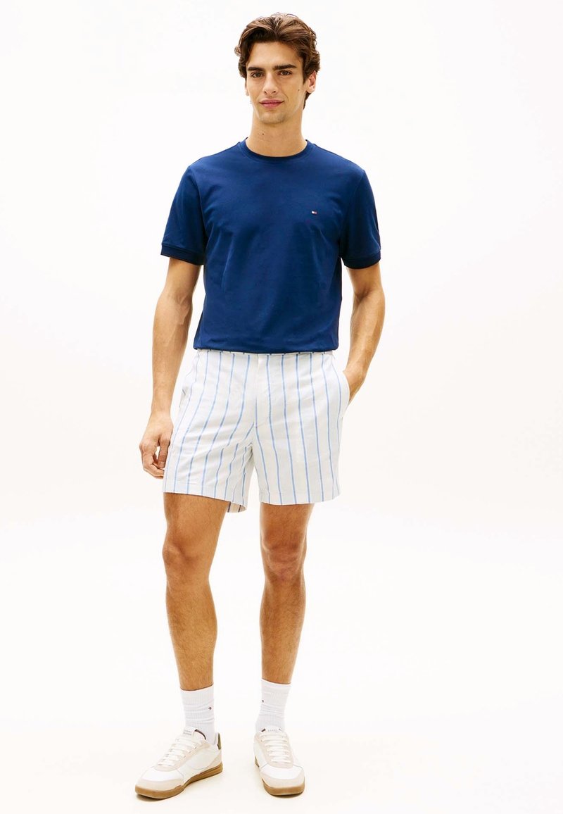 Jeune homme debout portant un t-shirt bleu marine, un short blanc avec des rayures verticales bleues, des chaussettes blanches et des baskets blanches, les mains dans les poches.