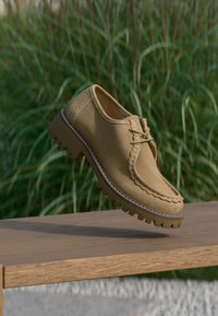 Chaussure beige en daim à lacets avec semelle épaisse et robuste survolant un banc en bois, sur fond d'herbe verte floue.