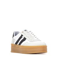 Zapatilla blanca con franjas negras y acentos grises. Presenta una suela de goma gruesa y cordones texturizados. Diseño de punta redondeada y tacón plano.