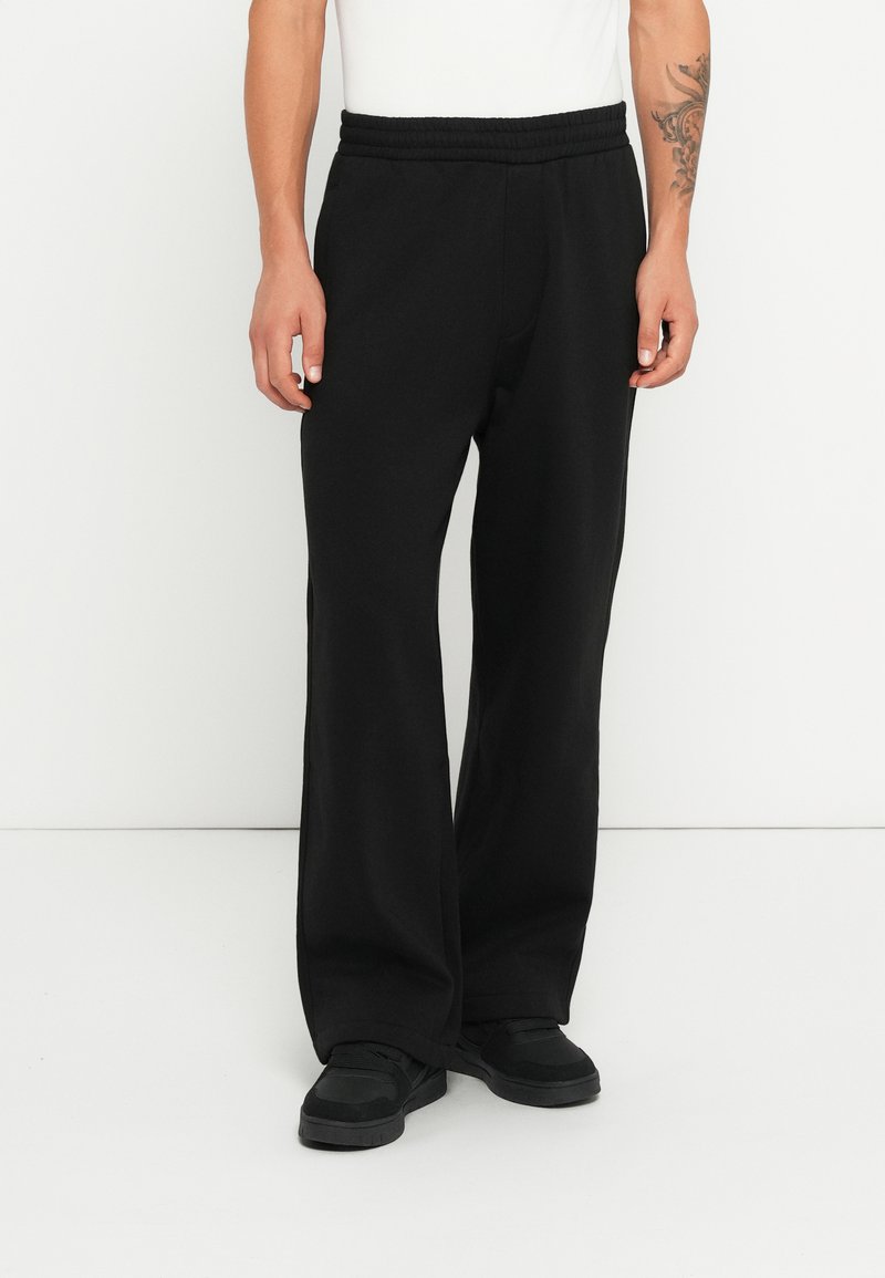 Schwarze Sweatpants mit einem elastischen Bund, die eine lockere Passform und Seitentaschen haben, kombiniert mit schwarzen Turnschuhen. Glatte Textur, einfaches Design.