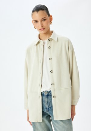 Lys beige jakke lavet af blødt stof, med kraveudskæring, knaplukning og løs pasform, parret med blå jeans.