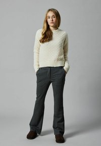 Cremfarbener gestrickter Pullover mit einem strukturierten Muster, kombiniert mit dunkelgrauen Nadelstreifenhosen. Das Model steht mit Händen in den Taschen.