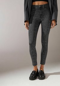 Jeans skinny de mezclilla negra con tachuelas metálicas plateadas, diseño de cintura alta y corte ajustado. Llevados con mocasines negros.
