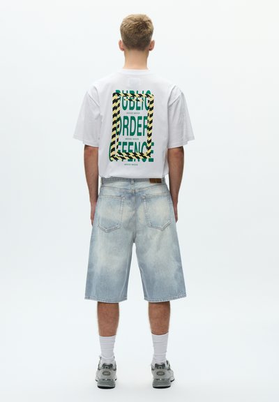 Fehér rövid ujjú póló zöld felirattal és fekete-sárga csíkos kerettel a hátoldalon. Világoskék farmer rövidnadrág látható zsebekkel és kifakult textúrával.