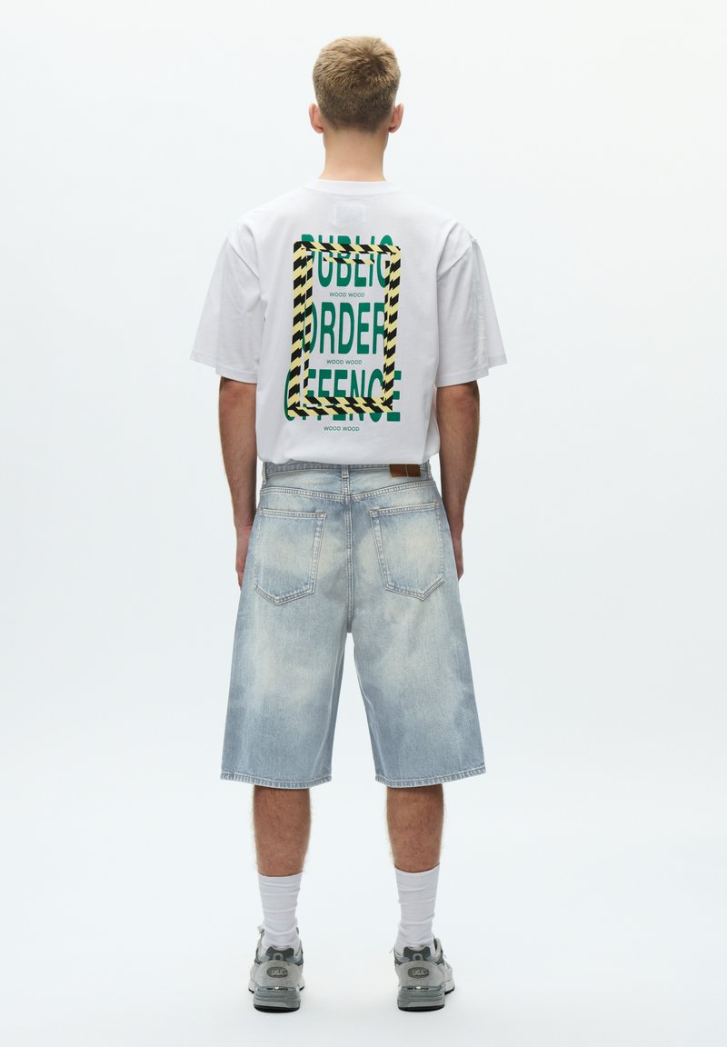 Weißes T-Shirt mit kurzen Ärmeln, grünem Text und schwarz-gelb gestreiftem Rahmen auf der Rückseite. Hellblaue Jeansshorts mit sichtbaren Taschen und verwaschenem Look.