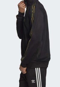 Giacca sportiva nera con alto colletto, dettagli a strisce mimetiche verdi sulle maniche, design aderente, realizzata in tessuto traspirante, abbinata a pantaloni coordinati.