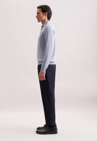 Mann in einem hellblauen Langarmpolohemd, dunklen marineblauen Hosen und schwarzen eleganten Schuhen, der im Profil vor einem schlichten Hintergrund steht.