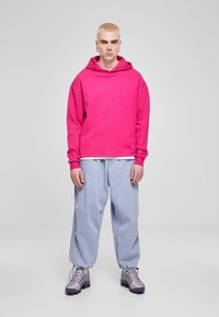 Sudadera fucsia con cordón y mangas ajustadas, combinada con unos vaqueros holgados de color azul claro y zapatillas grises con detalles texturizados.