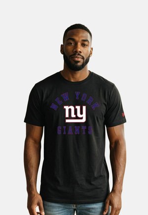 Man met kort haar en baard, gekleed in een zwart New York Giants T-shirt en blauwe jeans, staand voor een witte achtergrond.