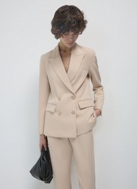 Beige Doppelreiher-Blazer mit schmalen Revers, zwei vorderen Taschen und passender Hose. Das Model hält eine schwarze, strukturierte Handtasche.