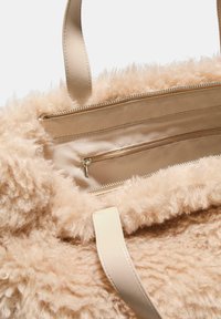 Bežová kabelka z umelého kožúška má vnútri hladkú podšívku, zipsový vrecko a kožené popruhy pre jednoduché nosenie. Mäkká textúra s plyšovým povrchom.