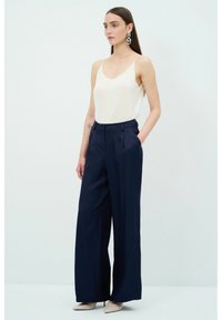 Marinblå vida byxor med framfickor och veck, kombinerat med en krämfärgad topp i camisole-stil. Modellen bär beiga klackar och iögonfallande örhängen.