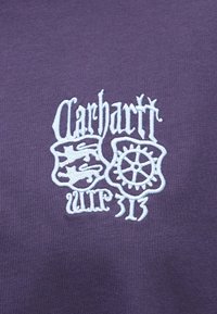 Fioletowa tkanina z białymi haftowanymi logo, w tym wzorem w kształcie koła zębatego oraz smoka, z dekoracyjnym tekstem i akcentami.