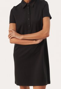 Donna che indossa un vestito polo nero a maniche corte con quattro bottoni, con le braccia incrociate, in piedi contro uno sfondo chiaro e semplice.
