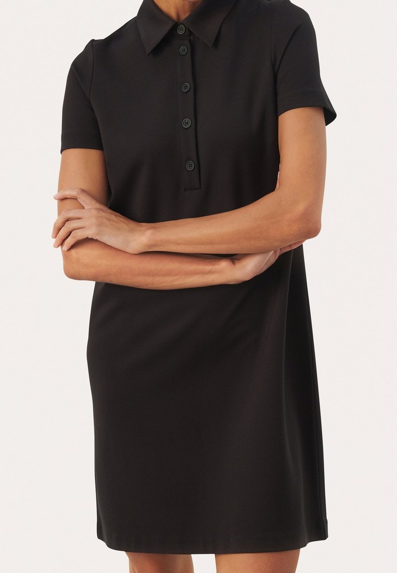 Donna che indossa un vestito polo nero a maniche corte con quattro bottoni, con le braccia incrociate, in piedi contro uno sfondo chiaro e semplice.