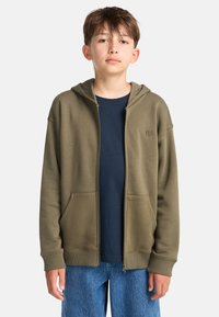 Ragazzo adolescente che indossa una felpa con zip verde oliva sopra una maglietta blu navy e jeans blu, in piedi contro uno sfondo grigio chiaro.
