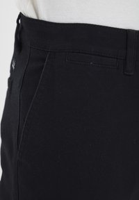 Pantalon en denim noir à texture lisse, présentant une coupe droite, des détails à la taille, et un logo discret près de la poche.